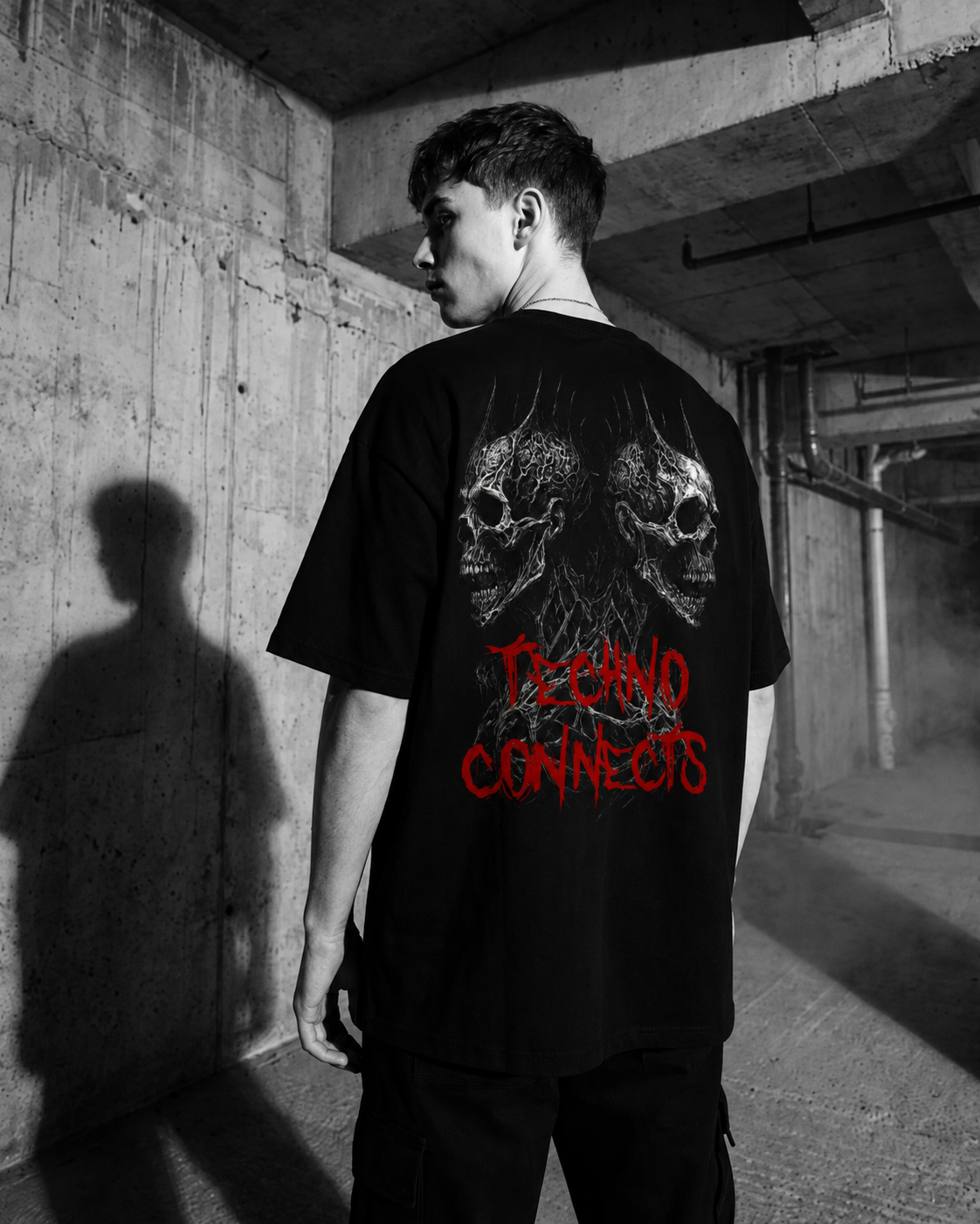 Model wearing the black Techno Connects oversized shirt, back view with skull graphic and red lettering, brutalist industrial rave photoshoot